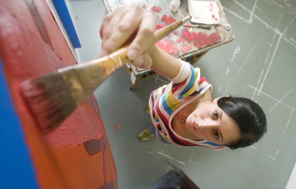 a looking down view of a student reaches high with her paint brush in red paint