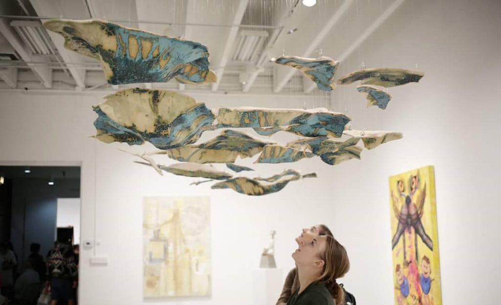 Student artwork by Diana Campuzano, two viewers looking up at the blue shatters of art work hanging from the ceiling