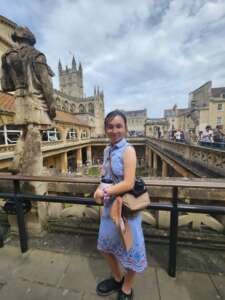 A smiling woman wearing a light-blue sleeveless dress and a beige cross-body bag leans on a wrought-iron railing overlooking the historic Roman Baths in Bath, England. Behind her you can see the honey-coloured Georgian stone buildings and the Gothic spires of Bath Abbey against a cloudy sky.